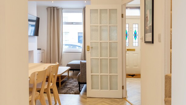 A room with a dining table and chairs, a sofa, a door with frosted glass panels, and a view of the street through a window. - Bright & Airy 2BD - 10min walk to Centre (Cheltenham)