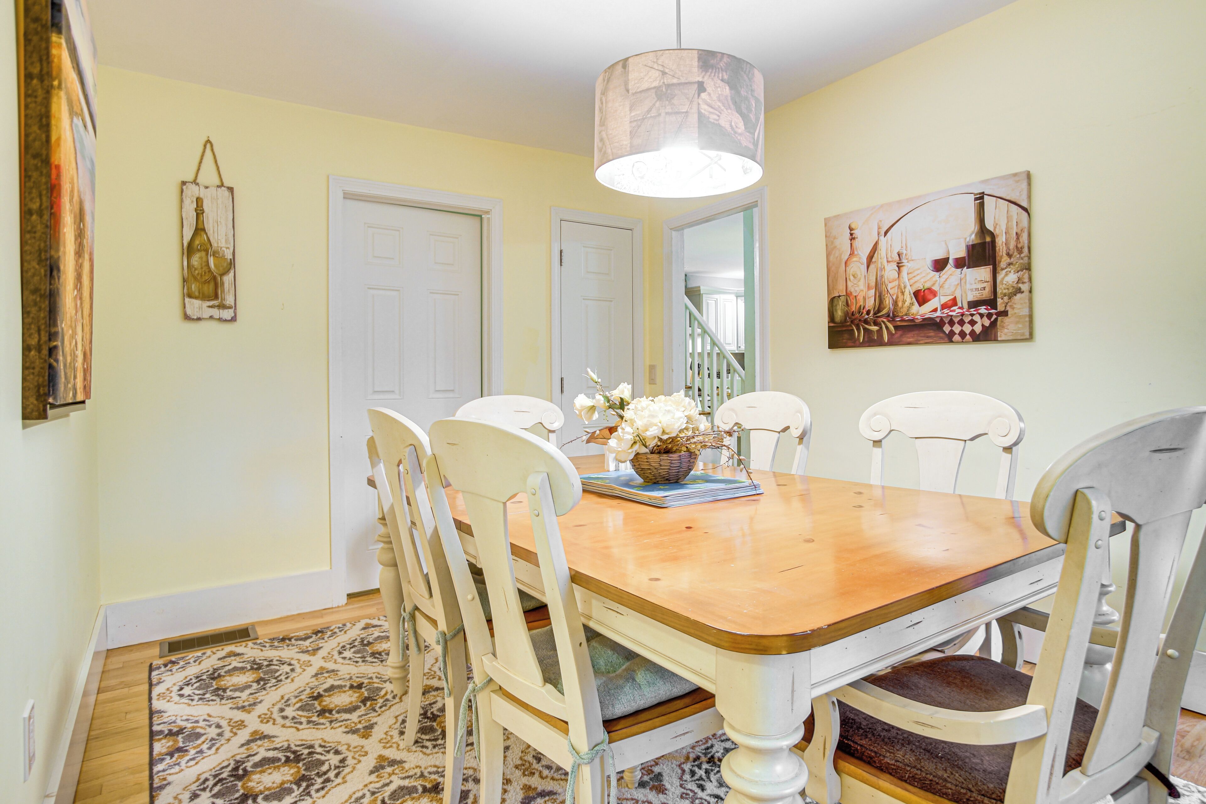 A dining room with a wooden table, white chairs, a hanging light fixture, and a painting on the wall.