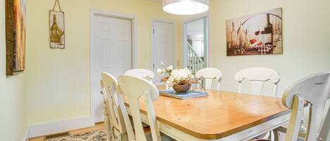 A dining room with a wooden table, white chairs, a hanging light fixture, and a painting on the wall.