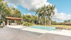 Una piscina amb un solàrium de pedra, una pèrgola de fusta i una zona verda amb palmeres.
