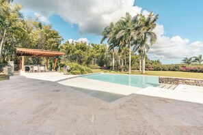 A swimming pool with a stone deck, a wooden pergola, and a grassy area with palm trees. - Apartment with 4 Bedrooms view in Casa de Campo (Casa De Campo)