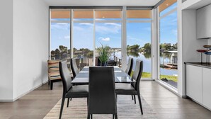 A modern dining area with a large table and chairs, large windows with a view of a lake and buildings, and a kitchen area with a white countertop and a red bowl.