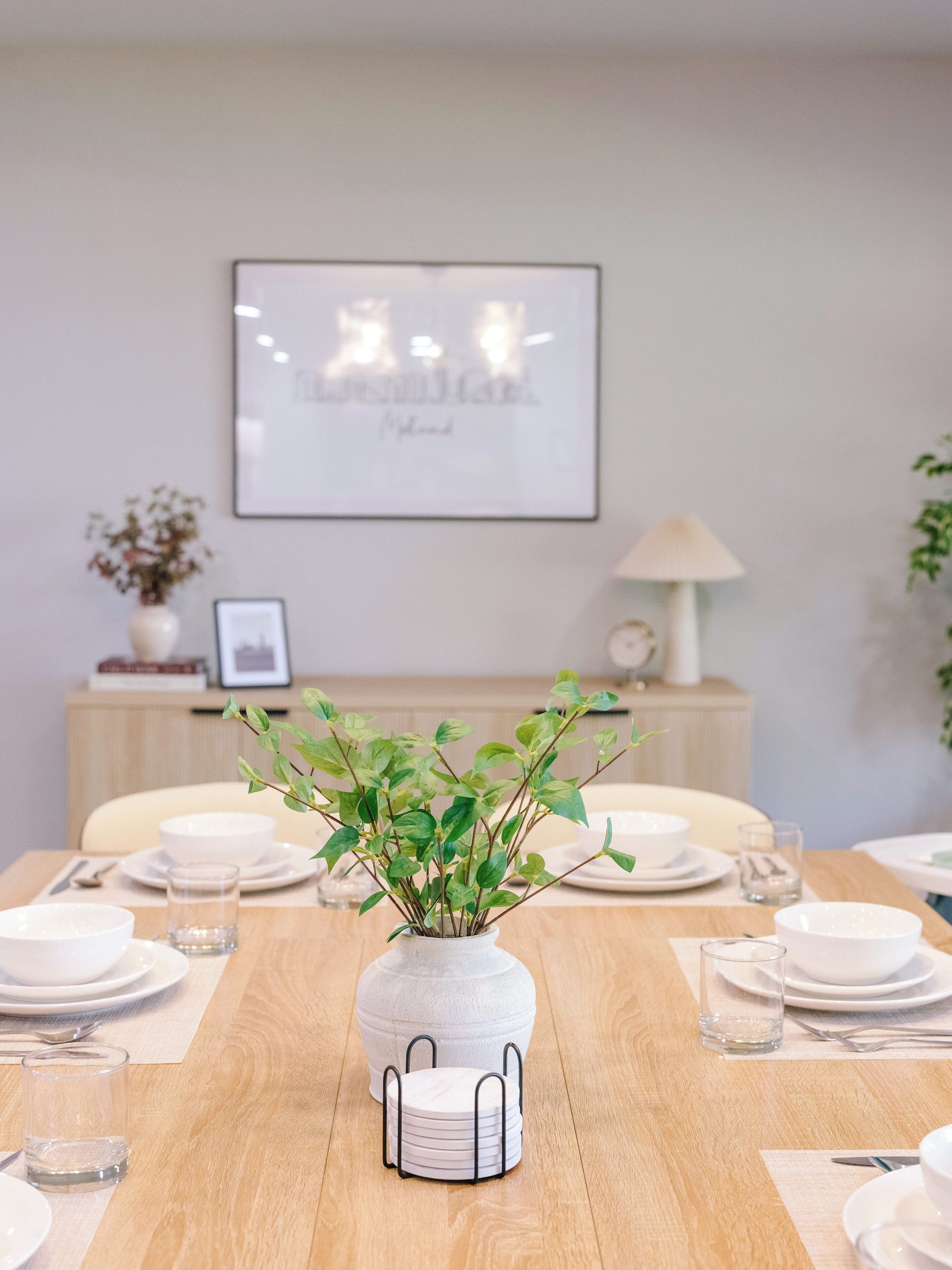 A dining table set with white plates, bowls, and glasses, centered with a white vase containing green plants.