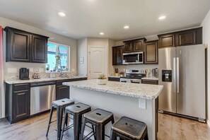 A modern kitchen with dark cabinetry, stainless steel appliances, a marble countertop, and a central island with stools. - Starlink Station! Cozy Retreat Near COS Airport (Colorado Springs)