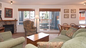 A living room with a stone fireplace, a patterned armchair, a green sofa, and a dining area with a wooden table and chairs. - Eagle Lodge corner unit 234 (Whistler)