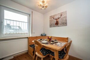 Uma área de refeições com uma mesa posta para quatro pessoas, um radiador abaixo da janela e uma pintura de um veado na parede.
