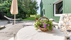 A patio with a table, a wicker basket with flowers, a lounge chair, and a green building in the background. - Apartment 'Deichblick' with Balcony and Wi-Fi (Wesselburenerkoog)
