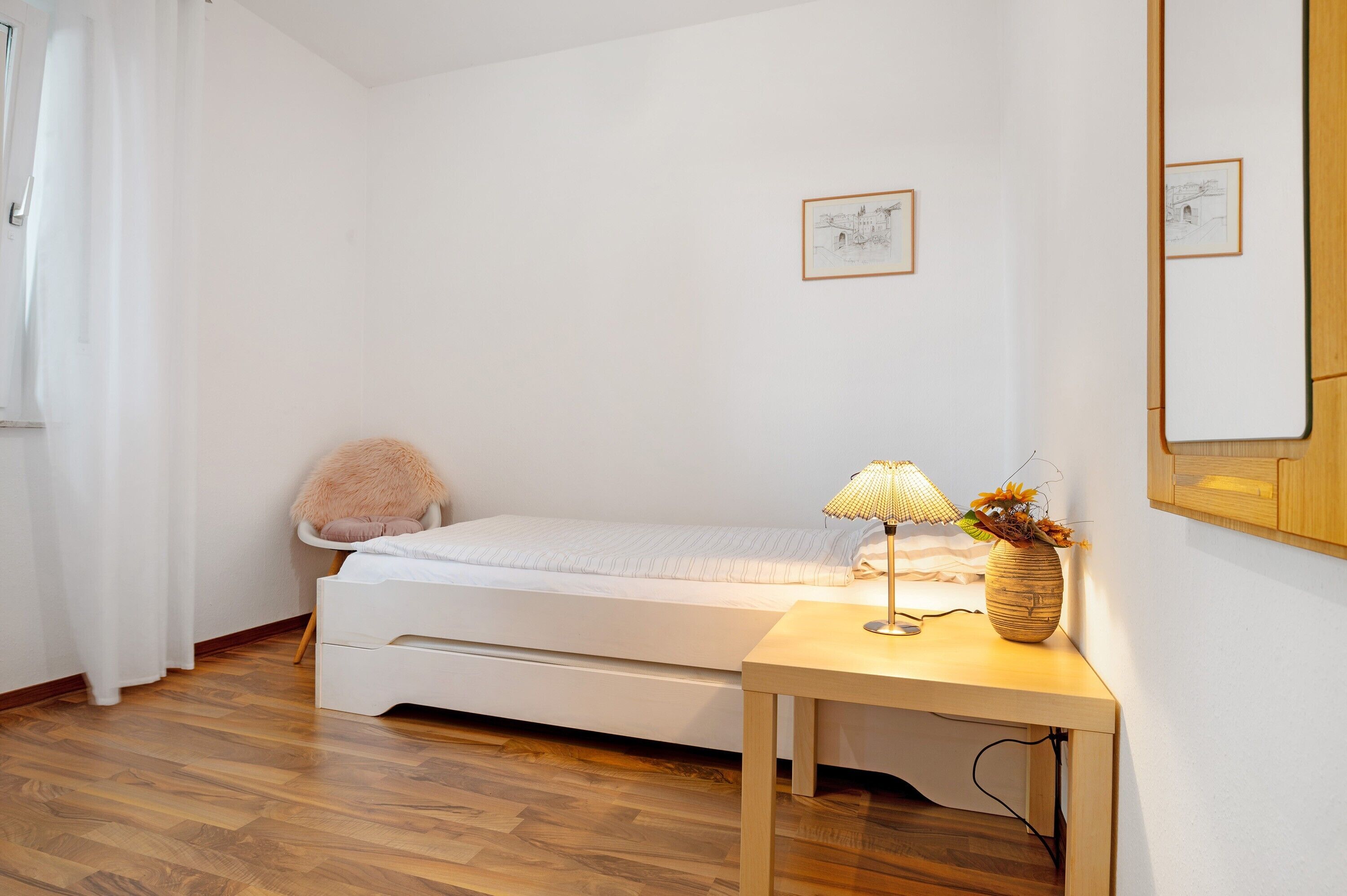 Een kleine, verzorgde slaapkamer met een enkel bed, een nachtkastje met een lamp en een vaas, en een spiegel aan de muur.