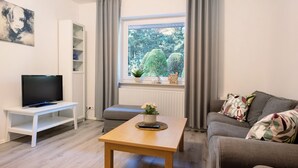 A living room with a TV on a white stand, a wooden coffee table, a grey sofa, and a window with a view of trees.