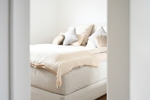 A bed with a beige blanket and pillows, a light-colored headboard, and a plain wall.