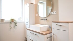 Ein Badezimmer mit einem weißen Waschbecken, einem Spiegel, einer Holzablage und einem Fenster mit einer Pflanze.