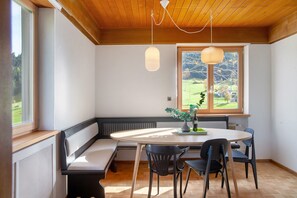 A room with a round table, four chairs, a bench, a window with a view of greenery, and a wooden ceiling.