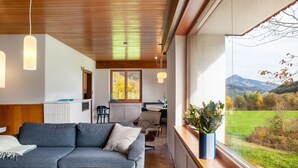 A modern living room with a sofa, a coffee table, pendant lights, and a view of a green landscape through large windows. - Holiday Home 'Blueberry Haus Im Moos' with Mountain View, Shared Terrace and Wi-Fi (Oberau)