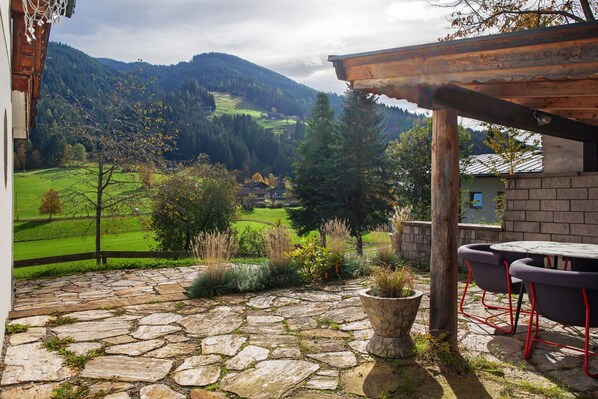 A stone patio with a wooden pergola, outdoor seating, and a view of a green valley and mountains.