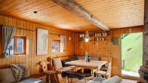 Interior de una cabaña de madera con una mesa de comedor, sillas, un sofá, chimenea y vistas al exterior.