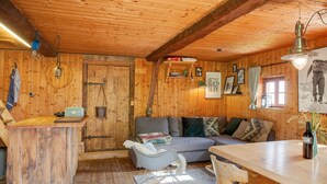 Interior de una cabaña de madera con sofá, mecedora, mesa de madera y una campana colgando en la pared.