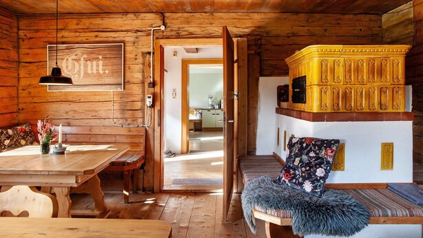 Interno di una baita in legno con tavolo, panche, divano e angolo cottura visibile attraverso una porta aperta.