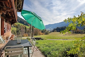 Une cabane en bois avec une table et des chaises à l’extérieur, un parasol vert et une vue sur une vallée verdoyante et des montagnes.