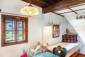 A room with a bed, a window with a view of greenery, a hanging light fixture, and a shelf with jars.