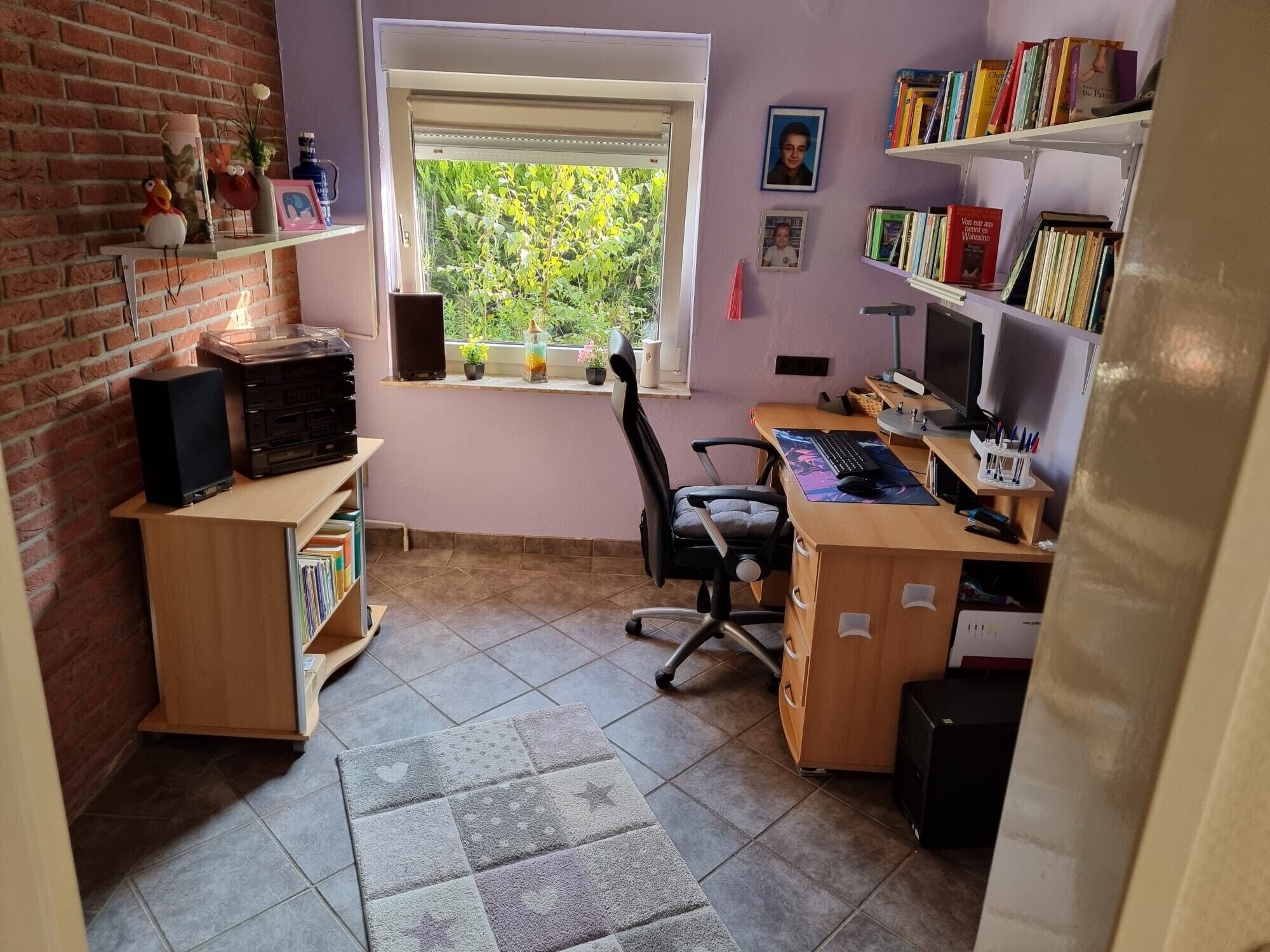 A room with a desk, chair, bookshelf, and a window with a view of greenery.