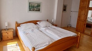 A wooden bed with a striped bedspread, a nightstand with a lamp, a painting on the wall, and a wooden wardrobe. - Apartment 'Ingrid Bauer 2' with Wi-Fi (Kühlungsborn)
