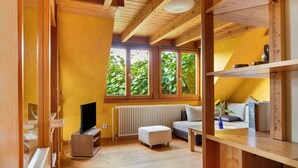 A room with wooden beams, a radiator, a television, a sofa, and a window with a view of greenery. - Apartment 'Fewo 1' with Wi-Fi (Müllheim im Markgräflerland)