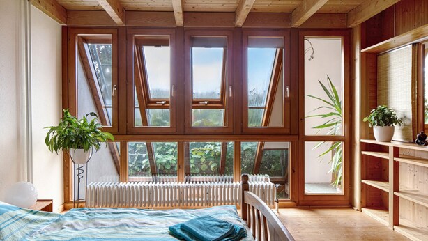A bedroom with a large window, wooden beams, a radiator, a bed with a blue blanket, and a potted plant. - Apartment 'Fewo 1' with Wi-Fi (Müllheim im Markgräflerland)