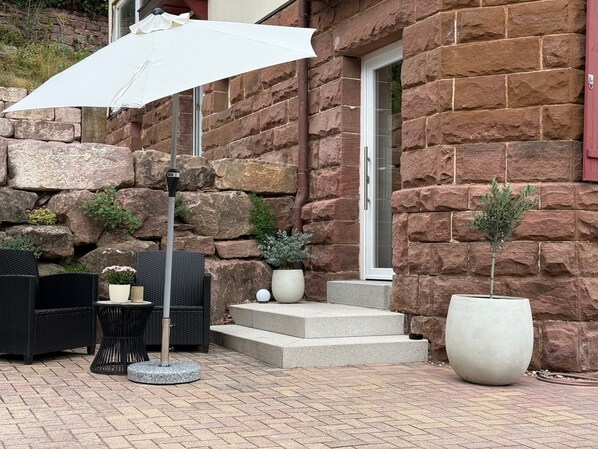 An outdoor seating area with wicker furniture, a white umbrella, and potted plants against a stone wall.