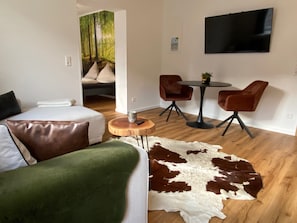 A modern living room with a sofa, a dining table with chairs, a flat-screen TV mounted on the wall, and a large painting of a forest scene.