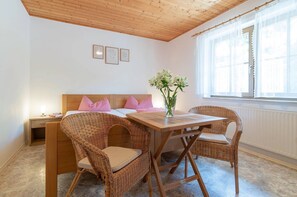 Una habitación con techo de madera, una cama con almohadas a cuadros, una mesa con sillas y una ventana con cortinas de gasa.