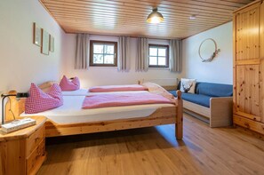 A room with two beds, a sofa, wooden floors, a ceiling with wooden beams, and a window with curtains.