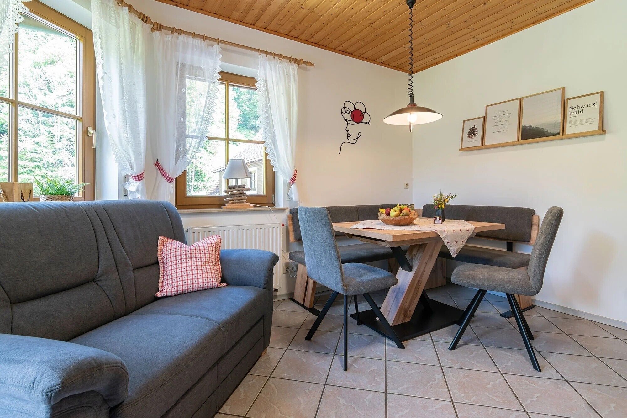 Ein gemütliches Zimmer mit einer grauen Couch, einem Esstisch mit Stühlen, einer Lampe und gerahmten Bildern an der Wand.