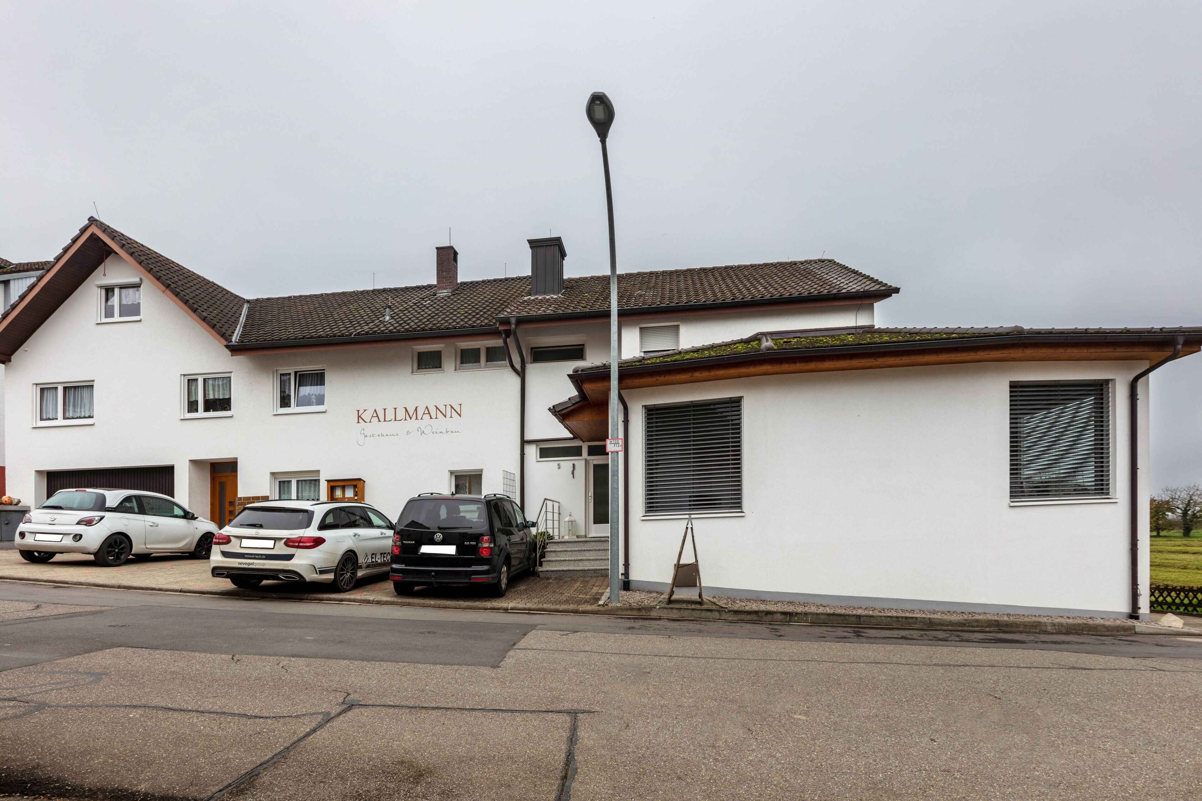 A building with a sign 'KALLMANN' and a parking area with cars.