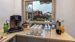 Una kitchenette con una cafetera, una bandeja de tazas blancas y una ventana con vistas a casas y vegetación.