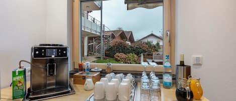 Una kitchenette con una cafetera, una bandeja de tazas blancas y una ventana con vistas a casas y vegetación.