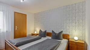 Cama doble con ropa de cama gris, cabecera de madera, dos mesitas de noche con lámparas, armario de madera y una puerta con cortina.