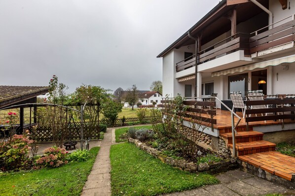 A residential building with a covered outdoor area, a garden with various plants, and a wooden staircase leading to a balcony.