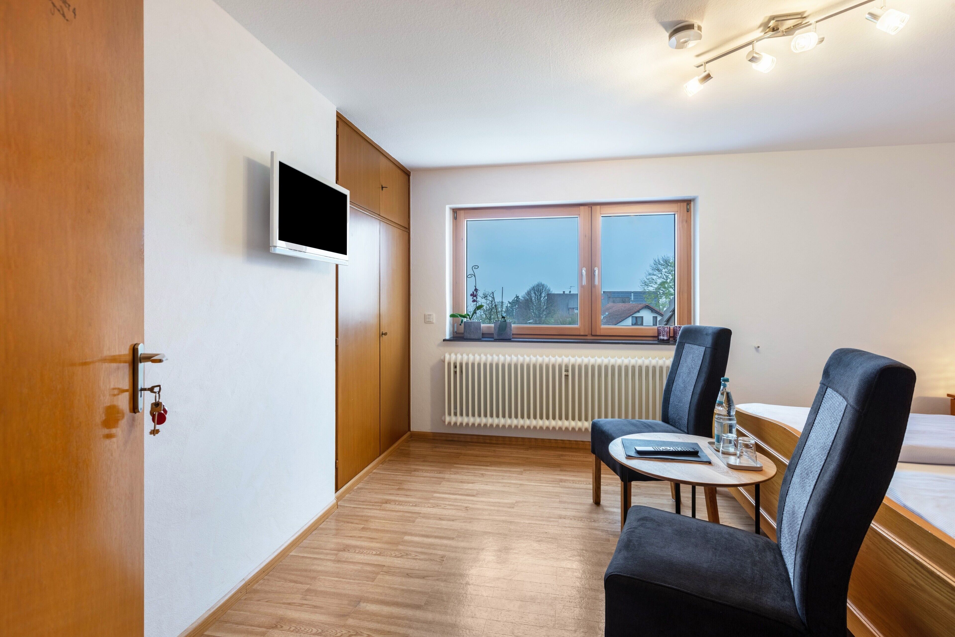 Ein Hotelzimmer mit Holztür, einem an der Wand befestigten Flachbildfernseher, einem Esstisch mit Stühlen, einem Fenster mit Blick auf Gebäude und einem Bett mit weißer Matratze.