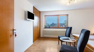 Ein Hotelzimmer mit Holztür, einem an der Wand befestigten Flachbildfernseher, einem Esstisch mit Stühlen, einem Fenster mit Blick auf Gebäude und einem Bett mit weißer Matratze.