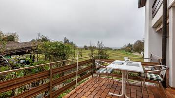 Un balcon avec une table et des chaises, une balustrade, et une vue sur un jardin et un paysage lointain.
