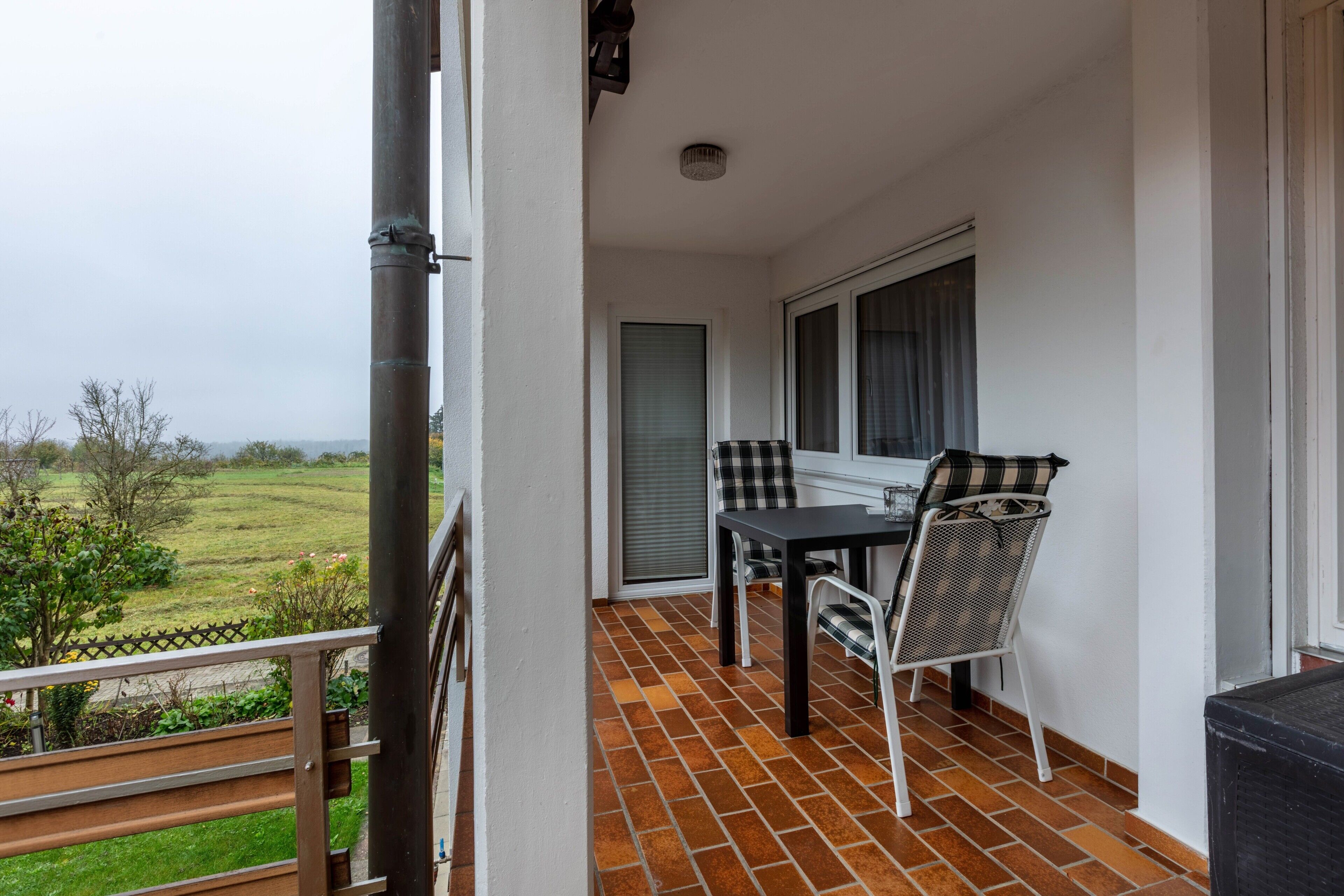 A balcony with a table and chairs, a window with a view of the countryside, and a door leading to the interior.