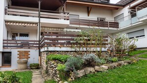 A multi-story building with balconies, a stone retaining wall, and a garden area with various plants.