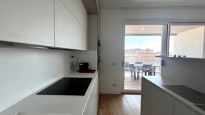 Une cuisine moderne avec des meubles blancs, un évier noir et une fenêtre donnant sur un balcon aménagé avec des sièges d’extérieur.