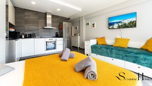 Habitación de hotel moderna con cocina, una cama con ropa de cama verde y amarilla, y un cuadro de una escena de playa.