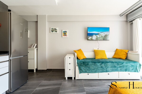A compact bedroom with a bed, a small bedside table, a wall-mounted TV, and a refrigerator.