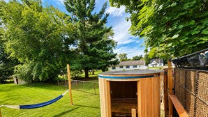 A wooden playhouse with a door, a hammock, and a fenced area with trees and a house in the background. - Vintage Rv & Spa Experience for 3 (Dover)