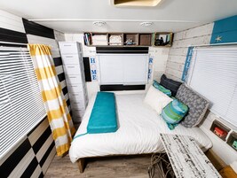 A compact bedroom with a bed, storage drawers, a window with blinds, and a small shelf with personal items.