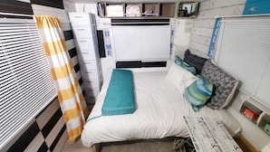 Un dormitorio pequeño con una cama, una ventana con persianas, una cortina y un estante con objetos.
