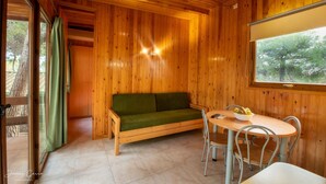 Interior de una cabaña de madera con sofá, mesa de comedor, sillas y una ventana que da a vista de árboles.