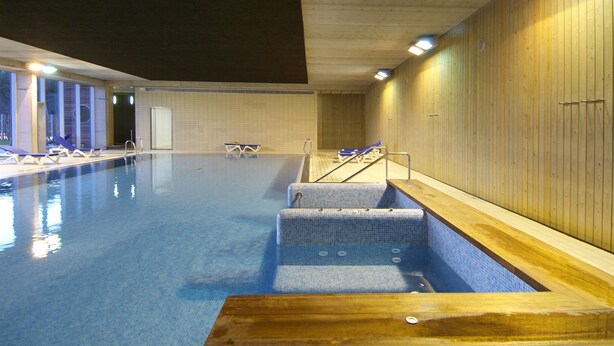 Una piscina interior con paredes de azulejos, una terraza de madera y sillones de descanso.